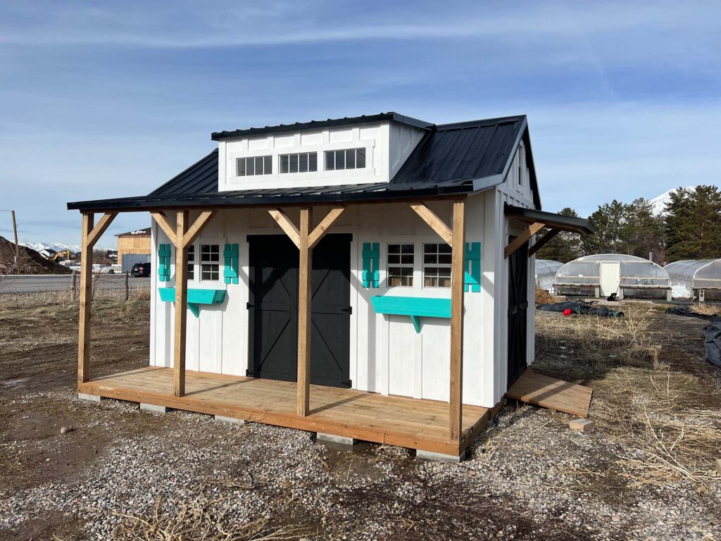 Custom garden shed with dormers, metal roof, covered porch, and deck built by Solitude Custom Sheds in Mapleton, Utah featuring white and turquoise designer paint colors.