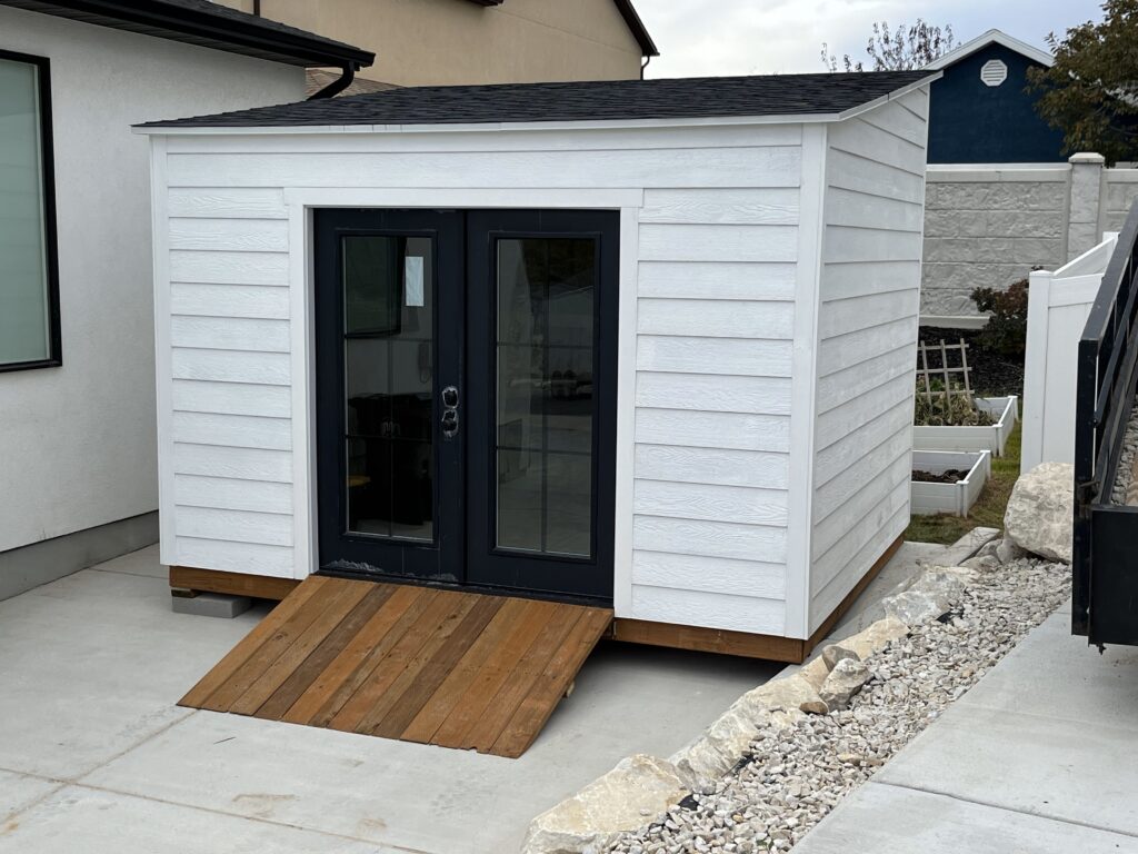 10x12 lean-to style backyard office shed with white horizontal lap siding, black grid French doors, and a custom wood ramp; built by Solitude Custom Sheds in Salt Lake City, Utah.