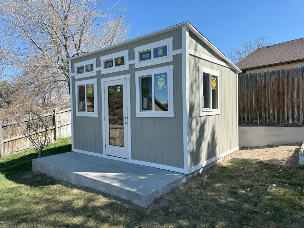 Modern 10x14 backyard home office shed in Sandy, Utah built by Solitude Custom Sheds featuring transom windows, and white trim on a concrete pad. Perfect set up for a backyard office.