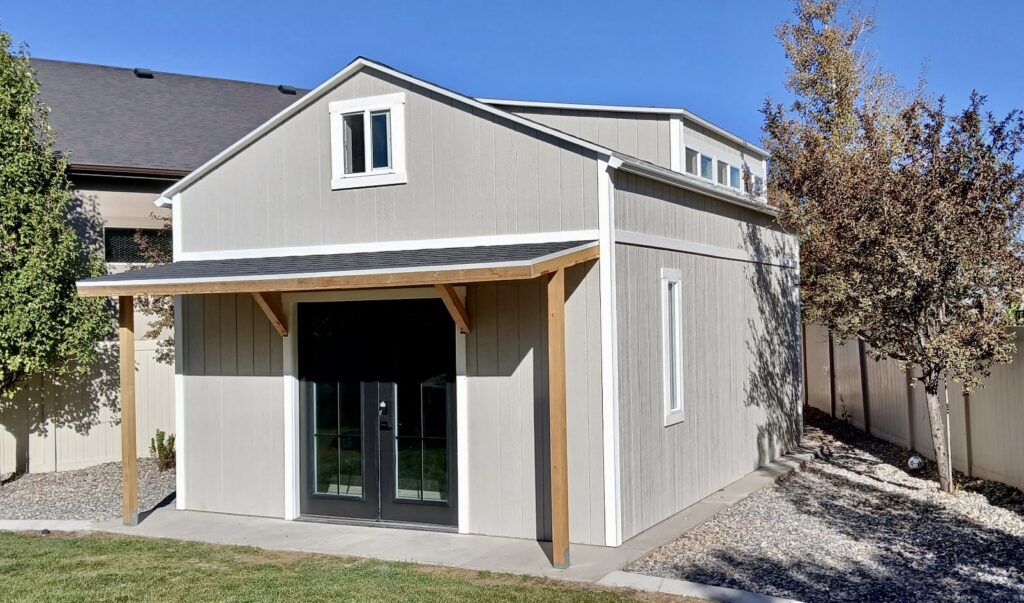 15x21 backyard golf simulator studio located in Herriman, UT and built by Solitude Custom Sheds; includes a modern gray exterior with white trim, black French doors, awning across the front, and frosted glass dormer windows. Perfect for home golf simulator studios, backyard offices, or backyard gyms.